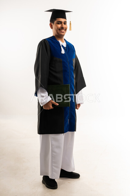 الوقوف باستعداد والنظر إلى الكاميرا بإيماءات السرور, بورتريه لخريج جامعي عربي خليجي عماني يرتدي الثوب والقبعه يمسك بيده مشروع التخرج, التعليم الاكاديمي في جامعات سلطنة عمان, مفهوم الدراسة والنجاح والتفوق, صورة الجسم بالطول الكامل, خلفية بيضاء