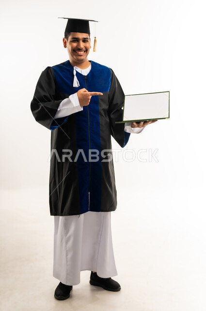 استخدام الأجهزة والتقنيات الحديثة والمتطورة, بورتريه لخريج جامعي عربي خليجي عماني يرتدي العباءة وقبعة التخرج يشير باصبعه إلى جهاز الحاسوب بشاشة فارغة, اظهار مشاعر السرور والسعادة, التعليم الاكاديمي في سلطنة عمان, صورة الجسم بالطول الكامل, خلفية بيضاء