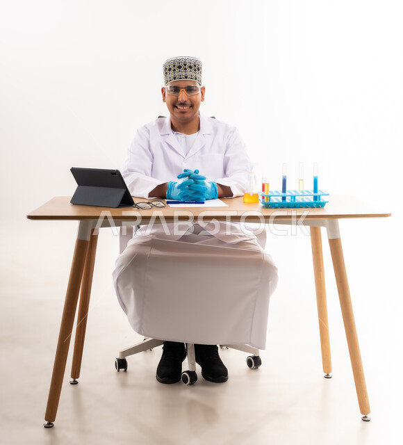 Academic education in the Sultanate of Oman, looking at the camera with a smile, chemical reactions inside the laboratory, conducting scientific experiments, using glass test tubes, portrait of an Arab Gulf Omani university student wearing a keffiyeh and a dishdasha sitting in a chemistry lab, white background