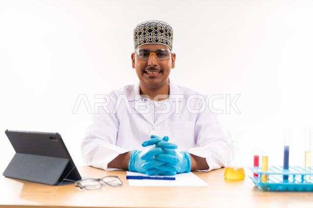 النظر إلى الكاميرا بابتسامة, التعليم الاكاديمي في سلطنة عمان, التفاعلات الكيميائية داخل المختبر, اجراء التجارب العلمية, اسخدام انابيب الاختبار الزجاجية, بورتريه مقرب لطالب جامعي عربي خليجي عماني يرتدي الكمه والدشداشه يجلس في معمل الكيمياء, خلفية بيضاء