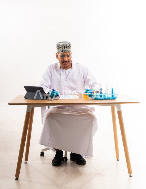 اجراء التجارب العلمية, التعليم الاكاديمي في سلطنة عمان, اسخدام انابيب الاختبار الزجاجية, دمج التعليم مع التقنية والتكنولوجيا, بورتريه مقرب لطالب جامعي عربي خليجي عماني يرتدي الكمه والدشداشه يقوم بتسجيل وتدوين نتائج التفاعلات الكيميائية في المختبر, خلفية بيضاء