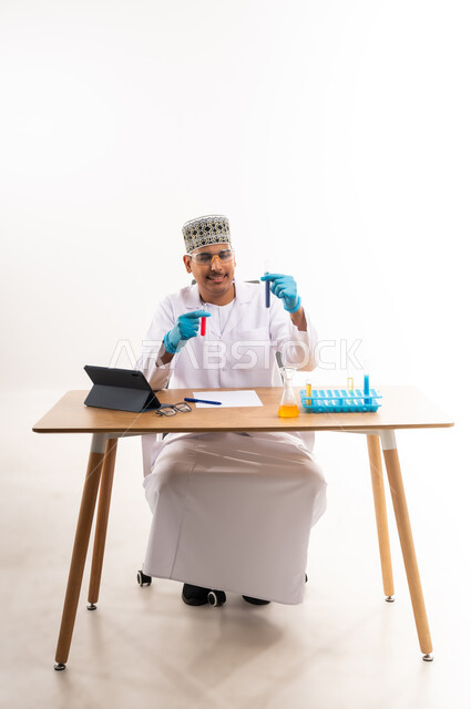 البحث عن نتائج التفاعلات الكيميائية, مفهوم الكيمياء الحيوية, بورتريه لطالب جامعي عربي خليجي عماني يرتدي الدشداشه والكمه يجلس في معمل الكيمياء, دمج التعليم مع التقنية والتكنوجيا, استخدام أنابيب الاختبار لإجراء التجارب في المختبر, خلفية بيضاء