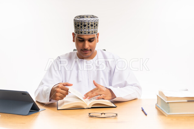 قراءة ومتابعة الدروس اليومية, بورتريه مقرب لطالب جامعي عربي خليجي عماني يرتدي الكمه والدشداشه يقوم بقراءة الكتاب, المناهج والمواد التعليمية, مفهوم التعليم الالكتروني في الجامعات الاكاديمية في سلطنة عمان, دمج التعلم مع التقنية والتكنولوجيا, خلفية بيضاء