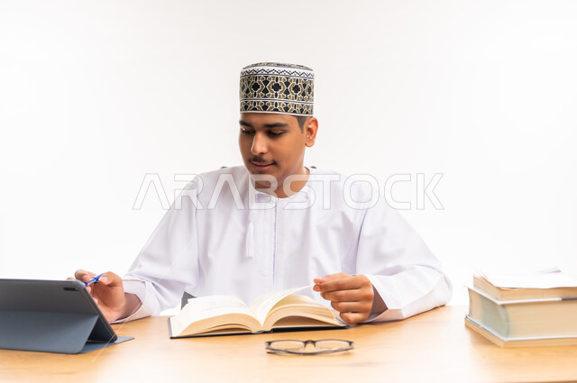 البحث الالكتروني عبر الانترنت, استخدام الأجهزة والتقنيات الحديثة والمتطورة, مفهوم التعليم عن بعد, بورتريه لطالب مقرب جامعي عربي خليجي عماني يرتدي الكمه والدشداشه يجلس على الطاولة ويعمل على جهاز التابليت, مراجعة ومتابعة الدروس والواجبات اليومية, خلفية بيضاء