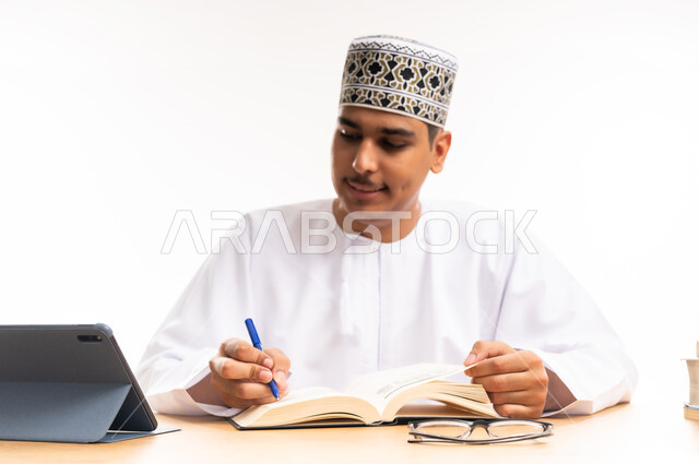 مراجعة الدروس اليومية, البحث الالكتروني عبر الانترنت, استخدام الأجهزة والتقنيات الحديثة والمتطورة, مفهوم التعليم عن بعد, بورتريه مقرب لطالب جامعي عربي خليجي عماني يرتدي الكمه والدشداشه يجلس على الطاولة ويقوم بتدوين المعلومات, خلفية بيضاء