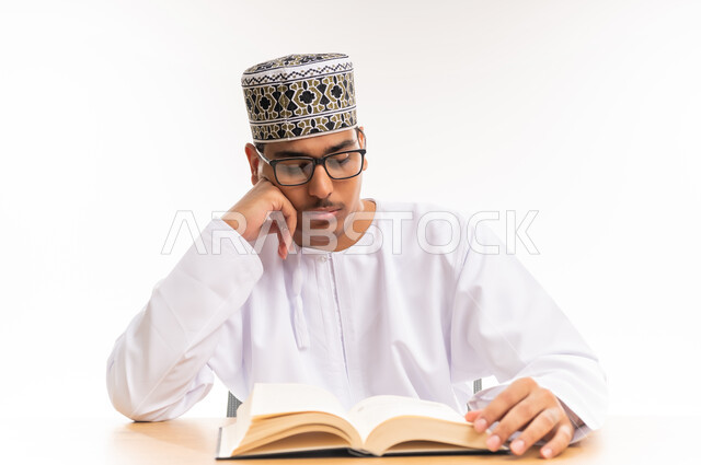 قضاء أوقات الفراغ بالأنشطة المفيدة, مفهوم التعلم والتفوق والاجتهاد, المناهج والمواد التعليمية في سلطنة عمان, الاستمتاع بممارسة هواية المطالعة, بورتريه مقرب لطالب جامعي عربي خليجي عماني يرتدي الدشداشه والكمه والنظارة الطبية يقوم بقراءة الكتاب, خلفية بيضاء