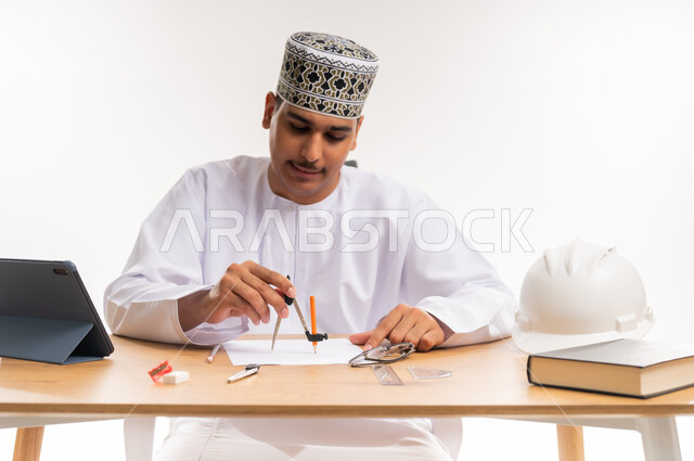 المناهج والمواد التعليمية, دمج التقنيات الحديثة والتكنولوجيا بالتعليم الأكاديمي, مفهوم الاجتهاد والتفوق, بورتريه مقرب لطالب جامعي عربي خليجي يرتدي الدشداشه والكمه يجلس على الطاولة في مكتبة الجامعة ويقوم بالرسم الهندسي باستخدام الفرجار, خلفية بيضاء