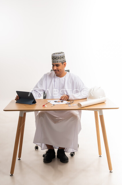 The concept of distance learning, smart electronic research, portrait of an Arab Gulf Omani university student wearing a dishdasha and a cloak sitting at a table and working on a tablet, the use of advanced devices and technologies in the educational field, curricula and educational materials in the Sultanate of Oman, white background