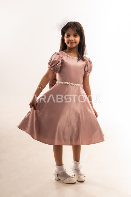 الاحتفال بالمناسبات والأعياد الدينية، الاهتمام بالجمال والمظهر الخارجي، فتاة عربية خليجية عمانية ترتدي لباس العيد الأنيق تمسك بيديها أطراف الفستان تنظر إلى الكاميرا بإيماءات السرور والفرح، فرحة وبهجة الأولاد، الجسم بالطول الكامل، خلفية بيضاء