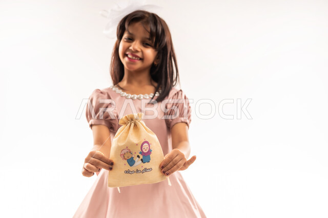 Bringing joy to the hearts of children, religious occasions and holidays, caring about beauty and appearance, a smiling Gulf Omani Arab girl wearing elegant Eid clothes holding a gift bag in her hands looking at the camera with gestures of joy and happiness, celebrating Eid al-Fitr, white background