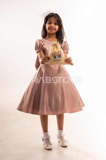 الاحتفال بالمناسبات والأعياد الدينية، الاهتمام بالجمال والمظهر الخارجي، فتاة عربية خليجية عمانية مبتسمة ترتدي لباس العيد الأنيق تمسك بيديها كيس هدايا تنظر إلى الكاميرا بإيماءات الفرح والسعادة، إدخال السرور على قلب الأبناء، الجسم بالطول الكامل، خلفية بيضاء