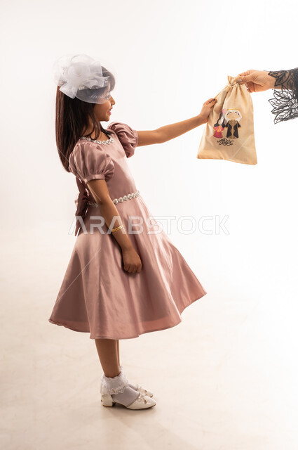 فرحة وبهجة الأولاد، الاهتمام بالجمال والمظهر الخارجي، صورة من الجانب لفتاة عربية خليجية عمانية ترتدي لباس العيد الأنيق تأخذ بيدها كيس هدايا، إدخال السرور والفرح على قلب الأبناء، الاحتفال بالمناسبات والأعياد الدينية، الجسم بالطول الكامل، خلفية بيضاء