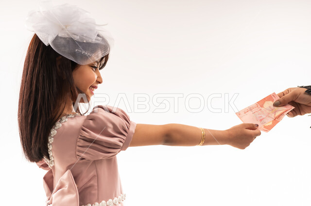 المناسبات والأعياد الدينية، الاهتمام بالجمال والمظهر الخارجي، تقديم العيدية للأبناء، يورتريه مقرب لفتاة عربية خليجية عمانية مبتسمة ترتدي لباس العيد الأنيق ترفع يدها تأخذ النقود، الاحتفال بعيد الفطر السعيد، خلفية بيضاء