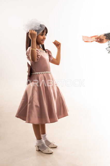 الاهتمام بالجمال والمظهر الخارجي، تقديم العيدية للأبناء، فتاة عربية خليجية عمانية مبتسمة ترتدي لباس العيد الأنيق تأخذ النقود ترفع يديها بإيماءات الفرح والحماس، المناسبات والأعياد الدينية، الاحتفال بعيد الفطر السعيد، الجسم بالطول الكامل، خلفية بيضاء