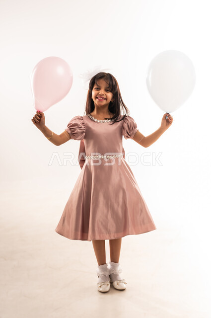 Religious occasions and holidays, celebrating Eid al-Fitr, smiling Omani Gulf Arab girl wearing elegant Eid dress holding colorful balloons in her hands looking at the camera with gestures of pleasure and happiness, interest in beauty and appearance, full body photo, white background