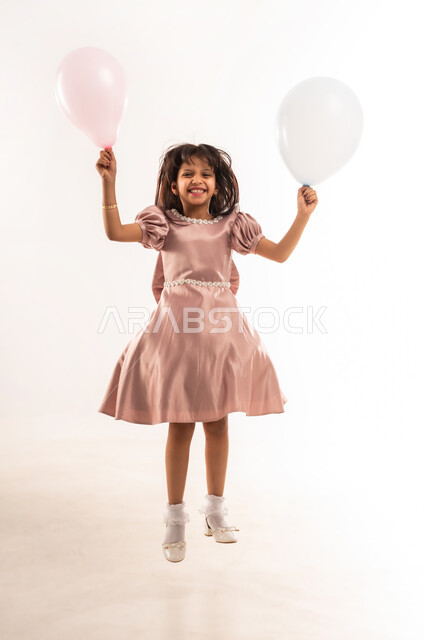 القفز عالياً تعبيراً عن الفرح والسرور، المناسبات والأعياد الدينية، الاحتفال بعيد الفطر السعيد، فتاة عربية خليجية عمانية مبتسمة ترتدي لباس العيد الأنيق تحمل بيديها بوالين ملونة تنظر إلى الكاميرا، الاهتمام بالجمال والمظهر الخارجي، الجسم بالطول الكامل، خلفية بيضاء