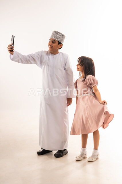 الاحتفال بالمناسبات الدينية، التواصل مع الأهل والأصدقاء وتبادل التهاني بمناسبة الأعياد، استخدام الأجهزة التقنية الحديثة، صبي وبنت عربيان خليجيان عمانيان يرتديان لباس العيد الأنيق يقومان بإجراء مكالمة فيديو عبر الهاتف المحمول، الجسم بالطول الكامل، خلفية بيضاء