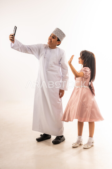 Communicating with family and friends and exchanging greetings on the occasion of the holidays, using modern technical devices, a girl raising her hand to greet, an Arab Gulf Omani boy and girl wearing elegant Eid clothes making a video call via mobile phone, full body, white background
