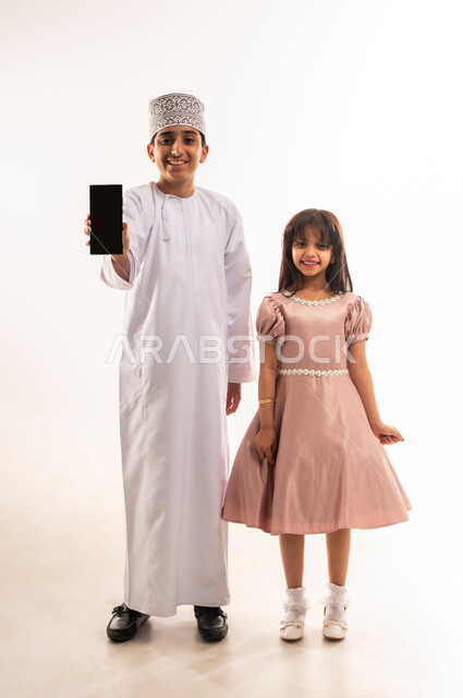 Using modern technology devices, having fun during the holidays, children's joy and happiness, boy showing a mobile phone with a black screen, two Arab Gulf Omani boy and girl wearing elegant Eid clothes looking at the camera with gestures of joy and happiness, full body, white background
