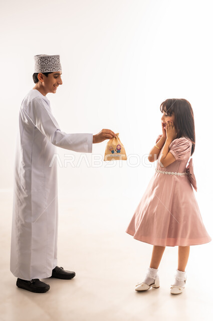 قضاء الأوقات الممتعة في الأعياد، فرحة وبهجة الأولاد، تقديم الهدايا في المناسبات والأعياد، ولد يقدم كيس ورقي لفتاة تضع يدها على خدها بإيماءات الدهشة والسرور، صبي وبنت عربيان خليجيان عمانيان يرتديان لباس العيد الأنيق، الجسم بالطول الكامل، خلفية بيضاء