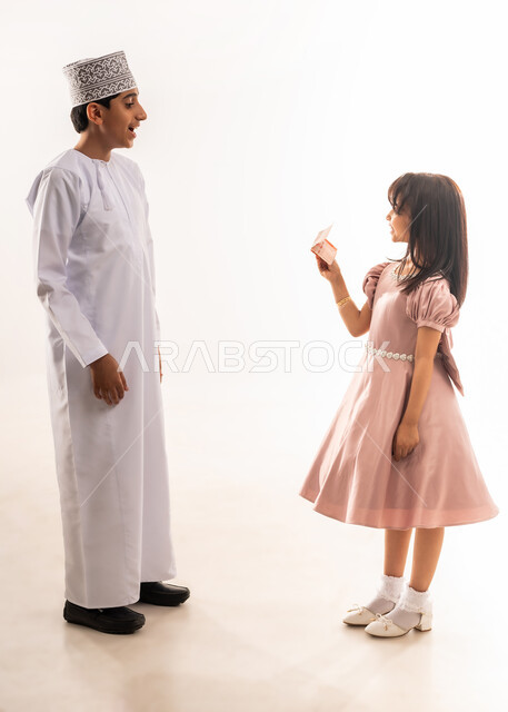 ولد ينظر بإيماءات الدهشة والسرور، المناسبات والأعياد الدينية، فناة تمسك بيدها نقود، تقديم العيدية للأبناء، صبي وبنت عربيان خليجيان عمانيان يرتديان لباس العيد الأنيق، قضاء الأوقات الممتعة في الأعياد، فرحة وبهجة الأولاد، الجسم بالطول الكامل، خلفية بيضاء