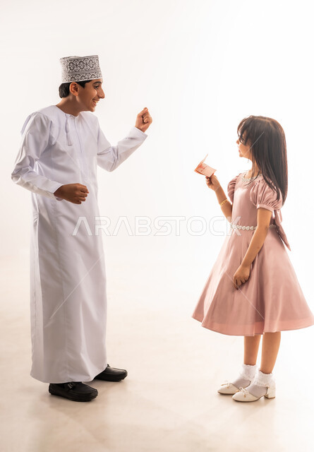 تقديم العيدية للأبناء، ولد يرفع يديه بإيماءات السرور والحماس، صبي وبنت عربيان خليجيان عمانيان يرتديان لباس العيد الأنيق، قضاء الأوقات الممتعة في الأعياد، فرحة وبهجة الأولاد، المناسبات والأعياد الدينية، فناة تمسك بيدها نقود، الجسم بالطول الكامل، خلفية بيضاء