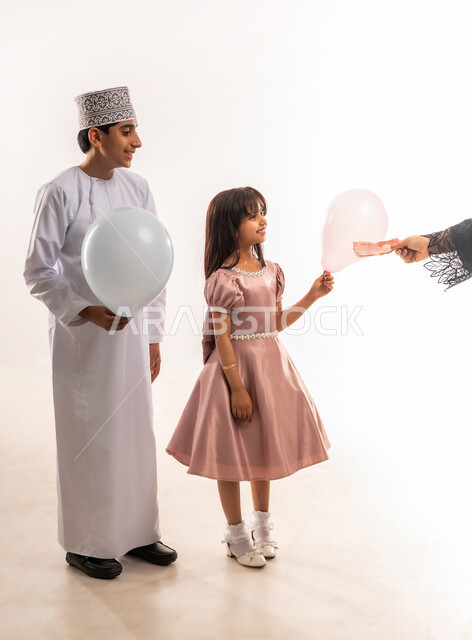 قضاء الأوقات الممتعة في الأعياد، تقديم العيدية للأبناء، الاحتفال بعيد الفطر السعيد، صبي وبنت عربيان خليجيان عمانيان يرتديان لباس العيد الأنيق يمسكان بيدهما بوالين بيضاء، فرحة وبهجة الأولاد، المناسبات والأعياد الدينية، الجسم بالطول الكامل، خلفية بيضاء