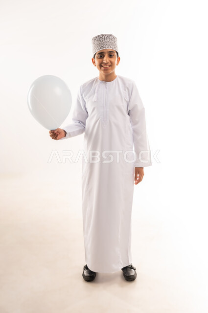 الاحتفال بعيد الفطر السعيد، صبي عربي خليجي عماني يرتدي الدشداشة و الكمة يحمل بيده بالون أبيض اللون ينظر إلى الكاميرا بإيماءات الفرح السعادة، قضاء الأوقات الممتعة في الأعياد، المناسبات والأعياد الدينية، الجسم بالطول الكامل، خلفية بيضاء