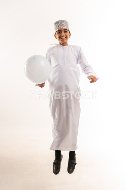 المناسبات والأعياد الدينية، الاحتفال بعيد الفطر السعيد، صبي عربي خليجي عماني يرتدي الدشداشة و الكمة يحمل بيده بالون أبيض اللون ينظر إلى الكاميرا بإيماءات السعادة، القفز عالياً تعبيراً عن الفرح والسرور، الجسم بالطول الكامل، خلفية بيضاء