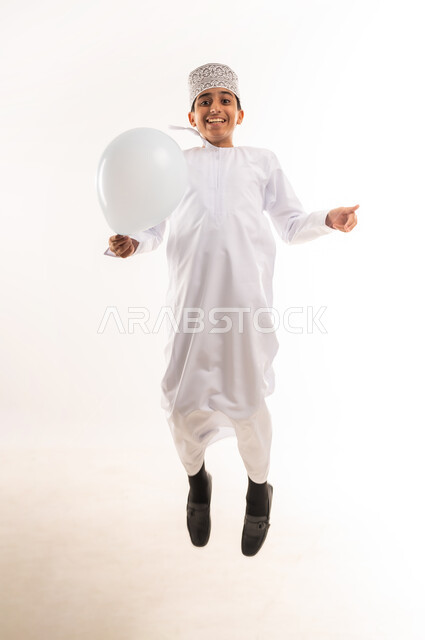 القفز عالياً تعبيراً عن الفرح والسرور، المناسبات والأعياد الدينية، الاحتفال بعيد الفطر السعيد، صبي عربي خليجي عماني يرتدي الدشداشة و الكمة يحمل بيده بالون أبيض اللون ينظر إلى الكاميرا بإيماءات السعادة، الجسم بالطول الكامل، خلفية بيضاء