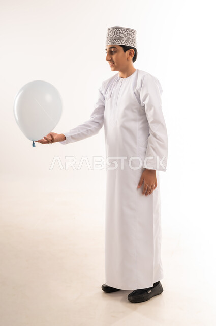 قضاء الأوقات الممتعة في الأعياد، الاحتفال بعيد الفطر السعيد، صبي عربي خليجي عماني يرتدي الدشداشة والكمة يحمل بيده بالون أبيض، فرحة وبهجة الأولاد، المناسبات والأعياد الدينية، الجسم بالطول الكامل، خلفية بيضاء