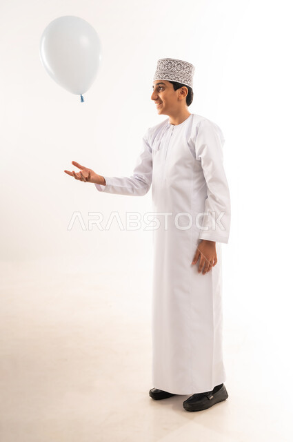 Children's joy and happiness, celebrating Eid al-Fitr, an Arab Gulf Omani boy wearing a dishdasha and a kimono playing with a white balloon, religious occasions and holidays, having fun on holidays, full body, white background