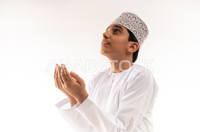 Striving in worship and getting closer to the Lord of the Worlds, close-up portrait of an Arab Gulf Omani boy wearing a dishdasha and a kummah raising his hands and praying to God with gestures of supplication and entreaty, desire to fulfill wishes, increasing supplication in the holy month of Ramadan, white background