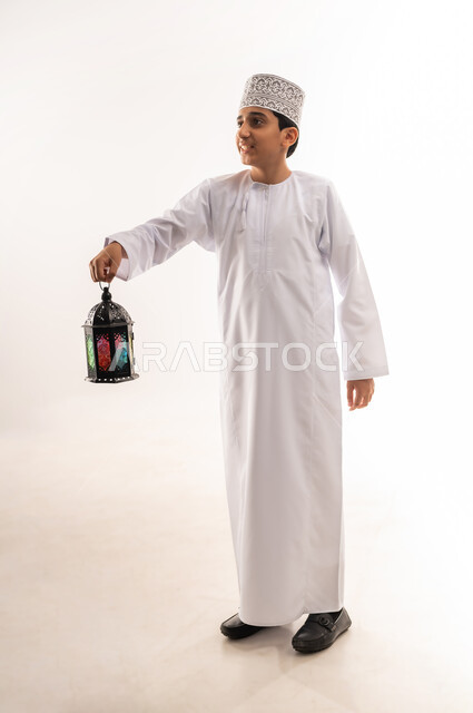 الاستعداد لاستقبال شهر رمضان المبارك بإيماءات السرور، فوانيس وزينة الأعياد والمناسبات الاسلامية في سلطنة عمان، صبي عربي خليجي عماني مبتسم يرتدي الدشداشة والكمة يمسك بيده فانوس ملون ينظر إلى شيء ما، صورة الجسم بالطول الكامل، خلفية بيضاء