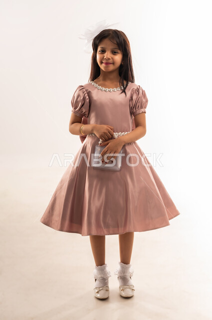 الاهتمام بالجمال والمظهر الخارجي، المناسبات والأعياد الدينية، فتاة عربية خليجية عمانية مبتسمة ترتدي لباس العيد الأنيق تمسك بيدها حقيبة صغيرة تنظر إلى الكاميرا بإيماءات السرور والسعادة، الاحتفال بعيد الفطر السعيد، الجسم بالطول الكامل، خلفية بيضاء