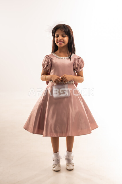 المناسبات والأعياد الدينية، فتاة عربية خليجية عمانية مبتسمة ترتدي لباس العيد الأنيق تمسك بيدها حقيبة صغيرة تنظر إلى الكاميرا بإيماءات السرور والسعادة، الاحتفال بعيد الفطر السعيد، الاهتمام بالجمال والمظهر الخارجي، الجسم بالطول الكامل، خلفية بيضاء