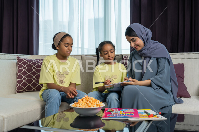 تنمية قدرات و مهارات الطفل, امراة عربية خليجية اماراتية مبتسمة ترتدي العباءة الكحلية تجلس على الاريكة تقوم بمساعدة ابنتيها في حل واجباتهن المنزلية, ايماءات السرور و الفرح, الديكورات والتصاميم الداخلية لغرف المعيشة في الفنادق في الامارات العربية المتحدة