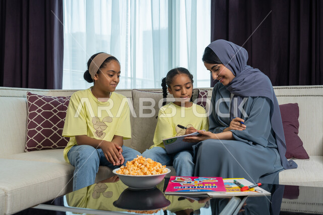التصاميم و الديكورات الداخلية لغرف المعيشة في الفنادق في الامارات العربية المتحدة, تنمية قدرات و مهارات الطفل, امراة عربية خليجية اماراتية مبتسمة ترتدي العباءة الكحلية تجلس على الاريكة تقوم بمساعدة ابنتيها في حل واجباتهن المنزلية, ايماءات السرور و الفرح