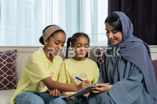 الديكورات والتصاميم الداخلية لغرف المعيشة في الفنادق في الامارات العربية المتحدة, تنمية قدرات و مهارات الطفل, امراة عربية خليجية اماراتية مبتسمة ترتدي العباءة الكحلية تجلس على الاريكة تقوم بمساعدة ابنتيها في حل واجباتهن المنزلية, ايماءات السرور و الفرح