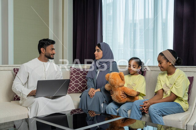 استخدام الاجهزة و التقنيات الحديثة المتطورة, ايماءات السرور و السعادة, رجل عربي خليجي اماراتي يرتدي الكندورة والغترة البيضاء يجلس في غرفة المعيشة يقوم بانجاز الاعمال الحاسب المحمول بجانب عائلته, الالفة والانسجام والمودة بين افراد الاسرة