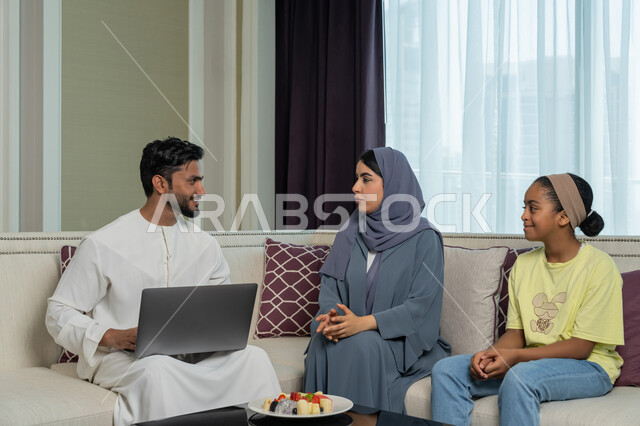 Using modern advanced devices and technologies, gestures of pleasure and happiness, an Emirati Arabian Gulf man wearing a white kandura and ghutra sitting in the living room completing work on a laptop next to his family, familiarity, harmony and affection among family members