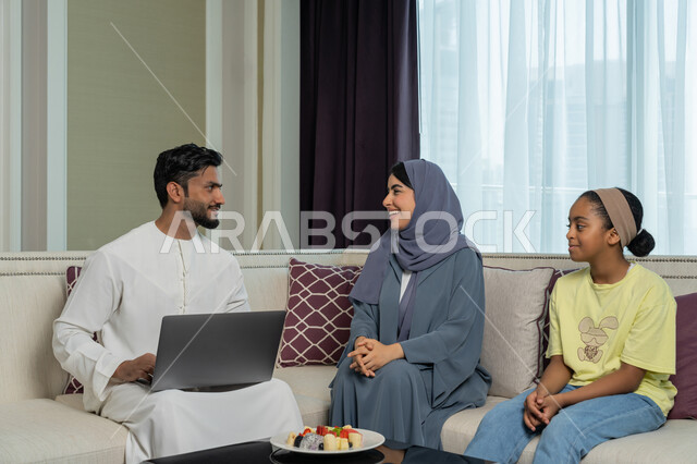 Using modern advanced devices and technologies, gestures of pleasure and happiness, an Emirati Arabian Gulf man wearing a white kandura and ghutra sitting in the living room completing work on a laptop next to his family, familiarity, harmony and affection among family members