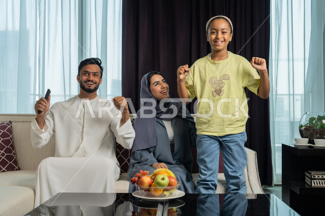 Enjoying spending time with family, raising the fist up with gestures of enthusiasm and joy for victory, an Arab Gulf Emirati family sitting in the living room watching the match on TV, passion to support the favorite football team, following the UAE national team