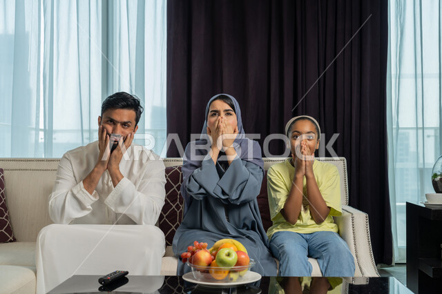 الاستمتاع بقضاء الأوقات الترفيهية الأسريه، روابط المحبه والتفاهم بين الأهل، رجل عربي خليجي اماراتي يرتدي الكندورة و الغترة البيضاء يجلس في غرفة المعيشة مع عائلته يضعون يديهم على فمهم, أسرة تشاهد التلفاز بايماءات تدل على الذهول و الصدمة