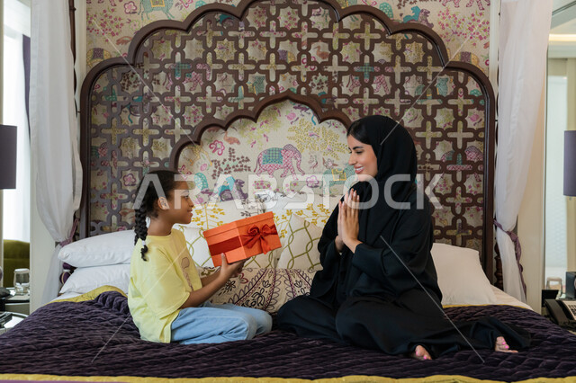 التعبير عن الحب والاحترام، تقديم الهدايا في المناسبات, إيماءات الفرح و السعادة، فتاة عربية خليجية اماراتية ترتدي الزي الكاجوال تجلس على السرير تمسك بيدها علبة هدية تقدمها لوالدتها بمناسبة عيد الام, الديكورات والتصاميم الداخلية لغرف النوم في الامارات