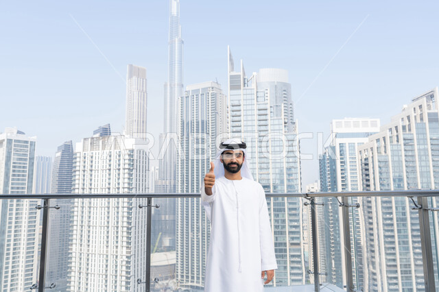 تعابير القبول والفرح والسرور، معالم سياحيه مشهورة في دبي، رجل عربي خليجي اماراتي مبتسم يرتدي الكندورة والغترة البيضاء يرفع اصبع السبابة للاعلى بإيماءات الإعجاب والرضا، مفهوم الاهتمام بالمظهر الخارجي، خلفية الأبراج وناطحات السحاب في الامارات العربية المتحدة