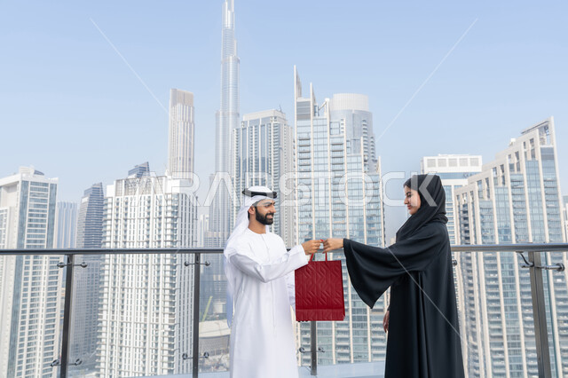 Celebrating occasions and holidays, urban growth and development, gestures of happiness and pleasure in enjoyable family gatherings in front of the Dubai Towers, expressing intimacy and love between spouses, an Arab Gulf Emirati man wearing a white kandura and ghutra presents his wife with a red bag on the occasion of Valentine's Day