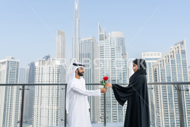 إيماءات السعادة والسرور بجلسات عائلية ممتعه امام ابراج دبي, التعبير عن الألفة والمحبة بين الزوجين, رجل عربي خليجي إماراتي يرتدي الكندورة و الغترة البيضاء يقدم لزوجته وردة حمراء بمناسبة عيد الحب, الاحتفال بالمناسبات و الأعياد, النمو والتطور العمراني، خلفية الأبراج وناطحات السحاب في الامارات العربية المتحدة