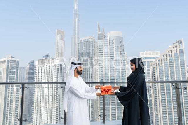 التعبير عن الألفة و المودة بين الزوجين, إيماءات السعادة والسرور, رجل عربي خليجي إماراتي يرتدي الكندورة و الغترة البيضاء يقدم لزوجته هديه في علبه حمراء بمناسبة عيد الحب, الاحتفال بالمناسبات و الأعياد، خلفية الأبراج وناطحات السحاب في الامارات العربية المتحدة