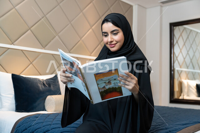 الاستمتاع بالأنشطة الترفيهية المفيدة في وقت الفراغ, مفهوم الثقافة والتعلم الذاتي, قراءة القصص والروايات, الإقامة في إحدى الفنادق الفاخرة، امراة عربية خليجية اماراتية ترتدي العباءة السوداء تجلس على سرير في غرفة النوم تحمل بيدها كتاب بإيماءات البشاشة والفرح